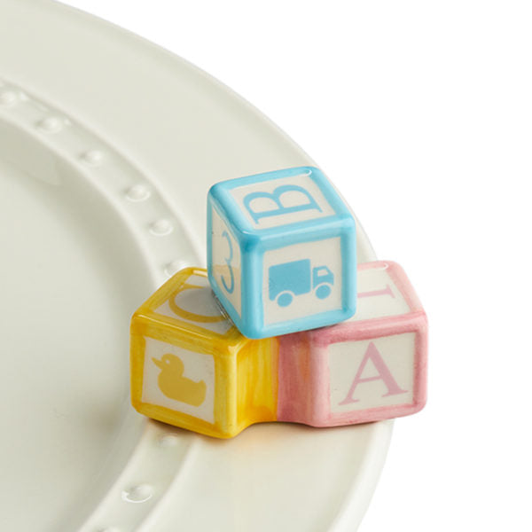 A porcelain trio of pastel blue, pink and yellow baby blocks sitting a top a Nora Fleming dish.