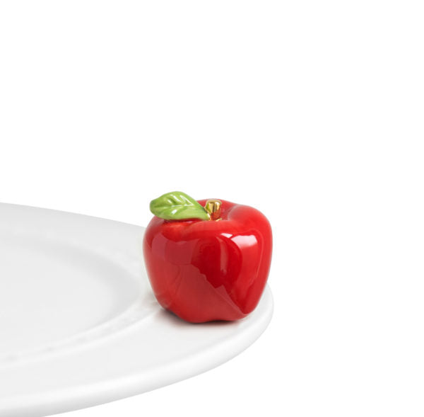 A shiny red apple with a green leaf ornament sitting a top a Nora Fleming dish.