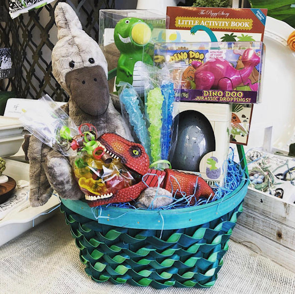 A green and blue woven basket filled with a plush dinosaur and other dinosaur related candies and gifts.